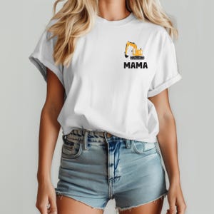 May include: A white short-sleeve t-shirt featuring a small graphic on the left chest area. The graphic depicts a yellow construction excavator above the black text MAMA. The shirt has a crew neckline and is paired with light blue denim shorts.