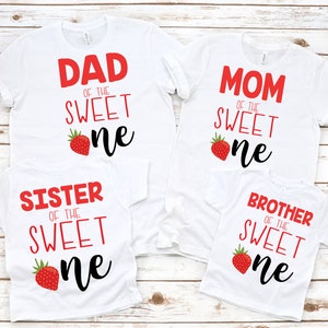 K&ouml;nnte beinhalten: Vier wei&szlig;e T-Shirts mit rotem Text und einer Erdbeergrafik. Die Shirts sagen "Papa vom s&uuml;&szlig;en Kleinen", "Mama vom s&uuml;&szlig;en Kleinen", "Schwester vom s&uuml;&szlig;en Kleinen" und "Bruder vom s&uuml;&szlig;en Kleinen".