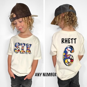 May include: Cream-colored t-shirt with the word "SIX" in a colorful comic book style on the front. The back of the shirt features the name "RHETT" and the number "6" in a similar design. The shirt is part of a set that can be customized with any number.