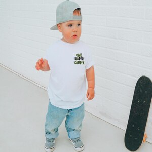 Puede incluir: Un ni&ntilde;o peque&ntilde;o con una camiseta blanca con el texto "ONE HAPPY CAMPER" en verde y negro. El ni&ntilde;o tambi&eacute;n lleva vaqueros azules, zapatillas grises y una gorra de b&eacute;isbol gris. Un monopat&iacute;n negro se apoya en una pared de ladrillos blancos.