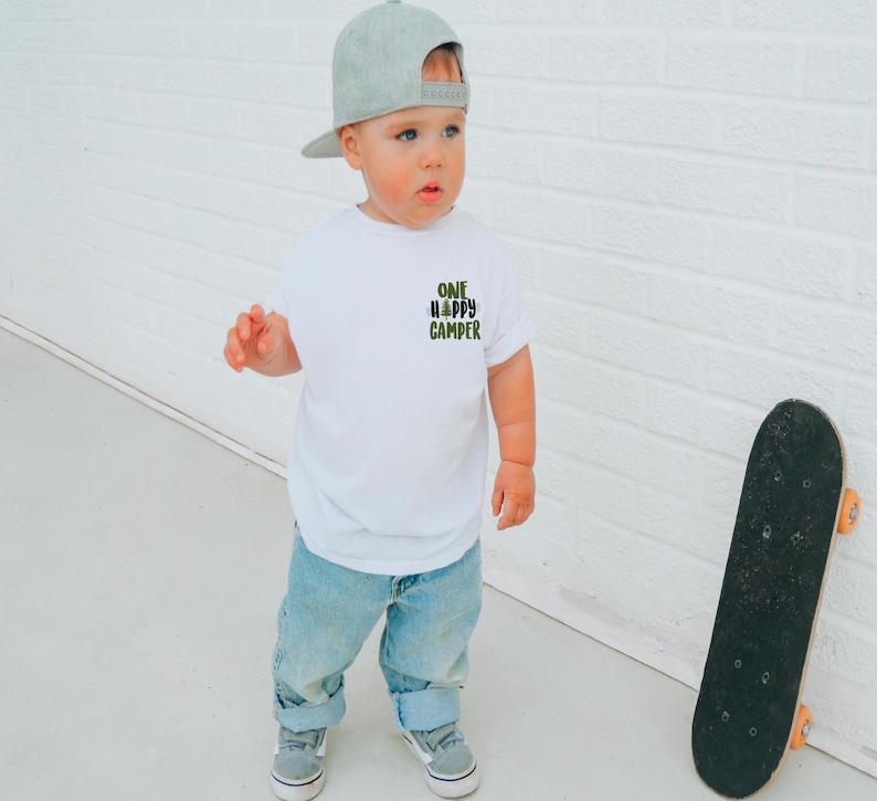 Puede incluir: Un ni&ntilde;o peque&ntilde;o con una camiseta blanca con el texto "ONE HAPPY CAMPER" en verde y negro. El ni&ntilde;o tambi&eacute;n lleva vaqueros azules, zapatillas grises y una gorra de b&eacute;isbol gris. Un monopat&iacute;n negro se apoya en una pared de ladrillos blancos.