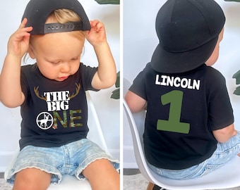 May include: A toddler wearing a black t-shirt with the text "The Big One" and a camouflage deer design. The toddler is also wearing a black baseball cap and denim shorts. The back of the shirt has the name "LINCOLN" and the number "1" in green.