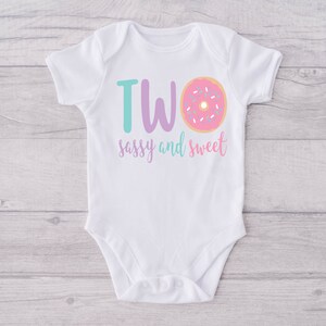 May include: White baby onesie with the text "TWO sassy and sweet" in purple and teal lettering. A pink donut with white sprinkles is next to the word "TWO".