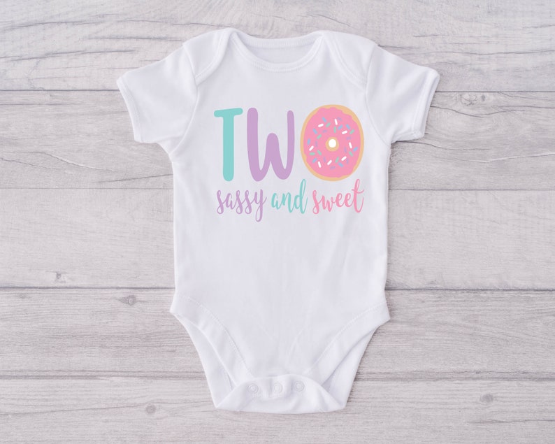 May include: White baby onesie with the text "TWO sassy and sweet" in purple and teal lettering. A pink donut with white sprinkles is next to the word "TWO".