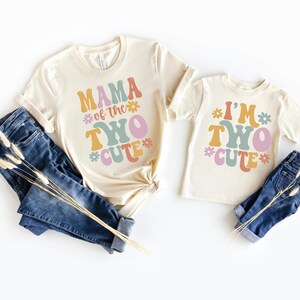 May include: Two cream-colored t-shirts with short sleeves and blue jeans. The larger shirt features the text MAMA of the TWO CUTE in colorful retro-style lettering with flower icons. The smaller shirt displays the text I'M TWO CUTE with flowers.