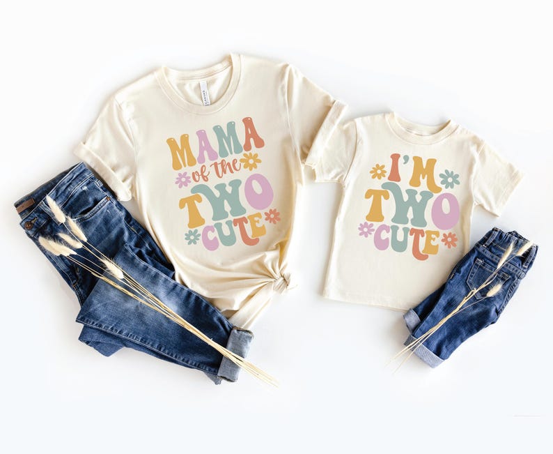 May include: Two cream-colored t-shirts with short sleeves and blue jeans. The larger shirt features the text MAMA of the TWO CUTE in colorful retro-style lettering with flower icons. The smaller shirt displays the text I'M TWO CUTE with flowers.