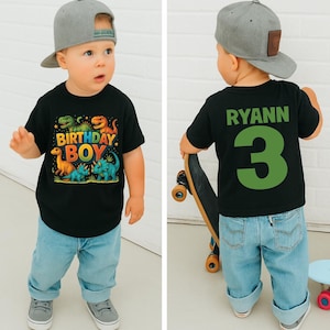 May include: A toddler wearing a black t-shirt with a colorful dinosaur graphic and the words "BIRTHDAY BOY." The back of the shirt has the name "RYANN" and the number "3" in green. The child is wearing a gray baseball cap and light blue jeans.
