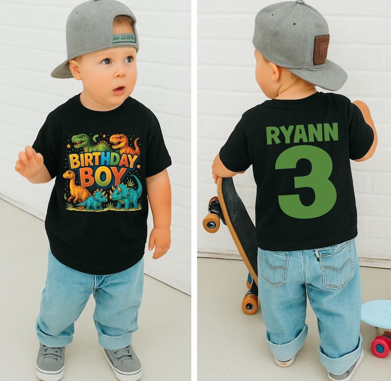 May include: A toddler wearing a black t-shirt with a colorful dinosaur graphic and the words "BIRTHDAY BOY." The back of the shirt has the name "RYANN" and the number "3" in green. The child is wearing a gray baseball cap and light blue jeans.