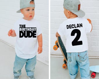 May include: A white t-shirt with black text that says "The Birthday Dude" on the front and "Declan 2" on the back. The shirt is being worn by a young boy wearing a grey baseball cap and blue jeans.