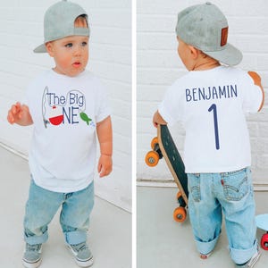 Op de afbeelding: Een peuter draagt een wit t-shirt met de tekst "The Big ONE" en een vissen-thema, gecombineerd met lichtblauwe jeans en een grijze baseballcap. De achterkant van het shirt heeft de naam "BENJAMIN" en het nummer "1".