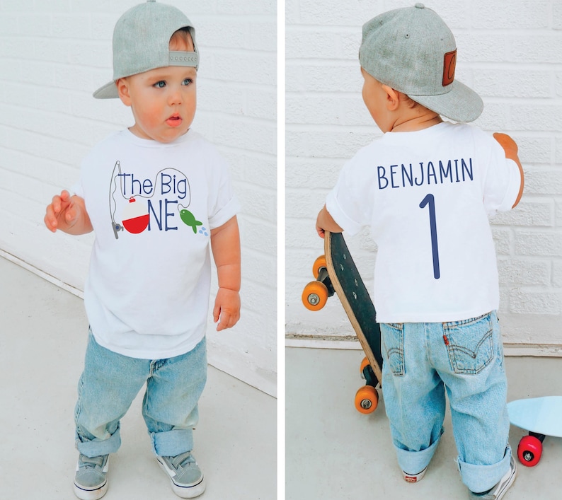Op de afbeelding: Een peuter draagt een wit t-shirt met de tekst "The Big ONE" en een vissen-thema, gecombineerd met lichtblauwe jeans en een grijze baseballcap. De achterkant van het shirt heeft de naam "BENJAMIN" en het nummer "1".
