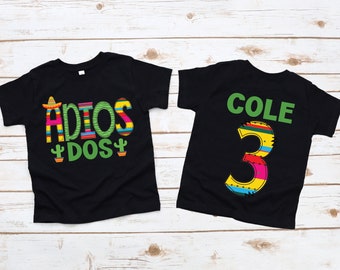 May include: Two black t-shirts with colorful designs. One shirt says "ADIOS DOS" with a sombrero and cactus. The other shirt says "COLE 3" with a colorful number three.