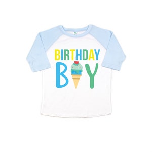 May include: White and light blue baseball tee shirt with a yellow, green, and blue graphic that says "Birthday Boy" with an ice cream cone in the middle.
