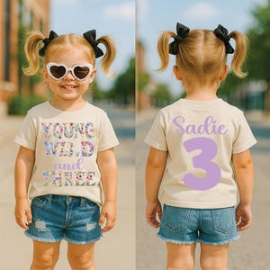 以下が含まれることがあります： フロントに花柄の「YOUNG WILD and THREE」の文字、バックにライトパープルの「Sadie」と数字の「3」がプリントされたベージュの半袖Tシャツ。ブルーのデニムショートパンツと黒いヘアリボンを合わせています。