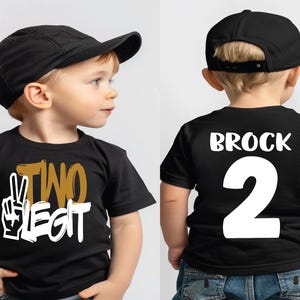 Pu&ograve; includere: Una maglietta nera con le parole "TWO LEGIT" in oro e bianco, e una grafica con il segno della pace. La parte posteriore della maglietta ha il nome "BROCK" e il numero "2" in bianco. Il bambino indossa un berretto da baseball nero.
