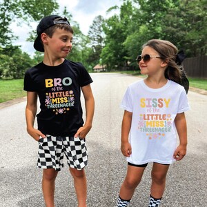 May include: A black t-shirt with the text BRO of the LITTLE MISS THREENAGER and a white t-shirt with the text SISSY of the LITTLE MISS THREENAGER, both featuring colorful floral graphics. The black shirt is paired with black and white checkered shorts.