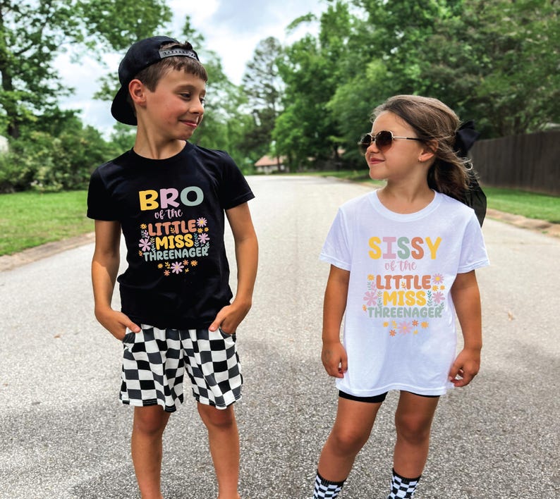 May include: A black t-shirt with the text BRO of the LITTLE MISS THREENAGER and a white t-shirt with the text SISSY of the LITTLE MISS THREENAGER, both featuring colorful floral graphics. The black shirt is paired with black and white checkered shorts.