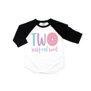 May include: A white and black baseball tee shirt with a pink donut and the text "TWO sassy and sweet".