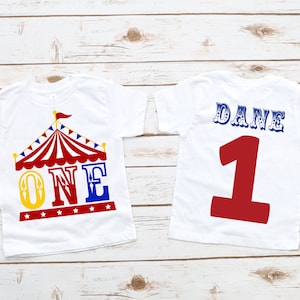 Puede incluir: Dos camisetas blancas con tem&aacute;tica de circo. La parte delantera de una camiseta tiene una carpa a rayas rojas y blancas con un "ONE" azul y amarillo debajo. La parte trasera de la otra camiseta tiene un n&uacute;mero "1" rojo con "DANE" azul y blanco encima.