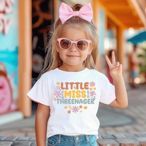 May include: A white short-sleeved t-shirt featuring the text Little Miss Threenager in orange, yellow, and green, surrounded by small colorful floral illustrations. The shirt is paired with blue denim jeans and a large light pink hair bow.