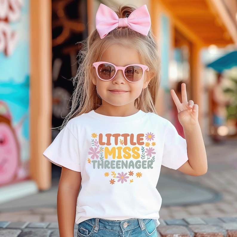 May include: A white short-sleeved t-shirt featuring the text Little Miss Threenager in orange, yellow, and green, surrounded by small colorful floral illustrations. The shirt is paired with blue denim jeans and a large light pink hair bow.