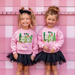 May include: Two girls wearing pink sweatshirts with green shamrock and leprechaun designs. The sweatshirts have the names "Emery" and "Lucia" on them. The girls are wearing black tulle skirts.