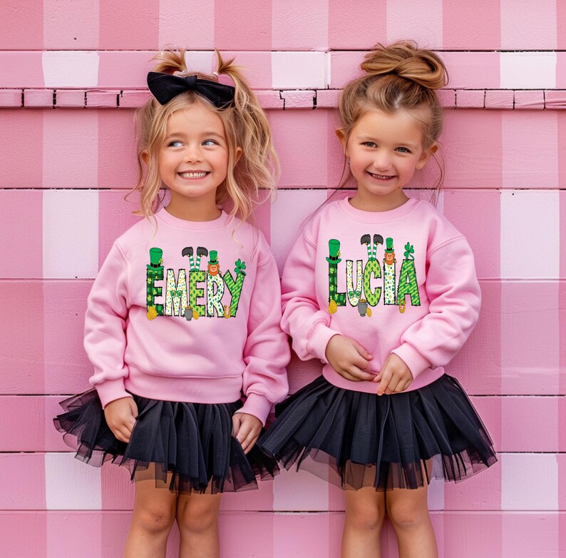 May include: Two girls wearing pink sweatshirts with green shamrock and leprechaun designs. The sweatshirts have the names "Emery" and "Lucia" on them. The girls are wearing black tulle skirts.