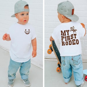 Op de afbeelding: Een peuter draagt een wit T-shirt met een hoefijzer en de naam "LUCAS" erop. De achterkant van het shirt toont "MY FIRST RODEO" met een afbeelding van een cowboy op een paard. De peuter draagt een blauwe spijkerbroek en een grijze baseball cap.