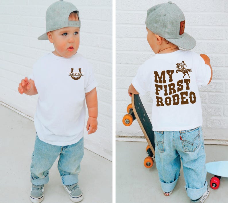 Op de afbeelding: Een peuter draagt een wit T-shirt met een hoefijzer en de naam "LUCAS" erop. De achterkant van het shirt toont "MY FIRST RODEO" met een afbeelding van een cowboy op een paard. De peuter draagt een blauwe spijkerbroek en een grijze baseball cap.