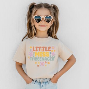 May include: A beige short-sleeved t-shirt featuring the text "LITTLE MISS THREENAGER" in orange, yellow, and green letters. The design is surrounded by small, colorful floral illustrations in shades of pink, yellow, and green.
