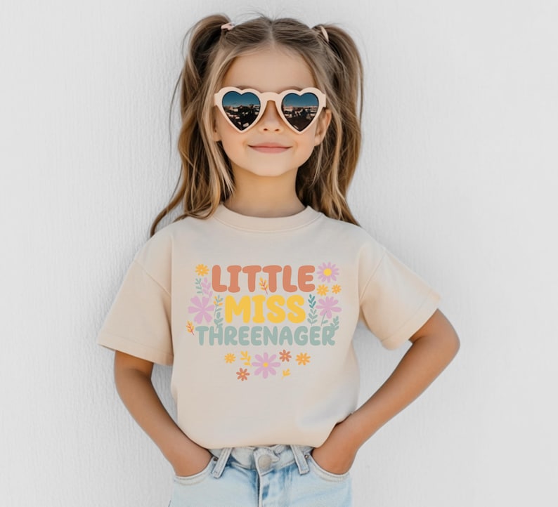 May include: A beige short-sleeved t-shirt featuring the text "LITTLE MISS THREENAGER" in orange, yellow, and green letters. The design is surrounded by small, colorful floral illustrations in shades of pink, yellow, and green.