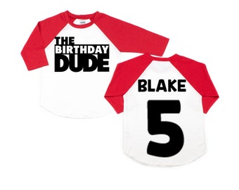 Może przedstawiać: Biało-czerwona koszulka w stylu bejsbolowym z czarnym napisem "The Birthday Dude" z przodu i "Blake 5" z tyłu.