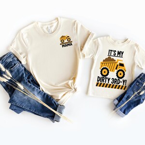 May include: Two cream-colored short-sleeve t-shirts and two pairs of blue denim jeans. One shirt features a small yellow dump truck graphic with the text MAMA. The other shirt features a large yellow dump truck graphic with the text IT'S MY DIRTY 3RD-Y!