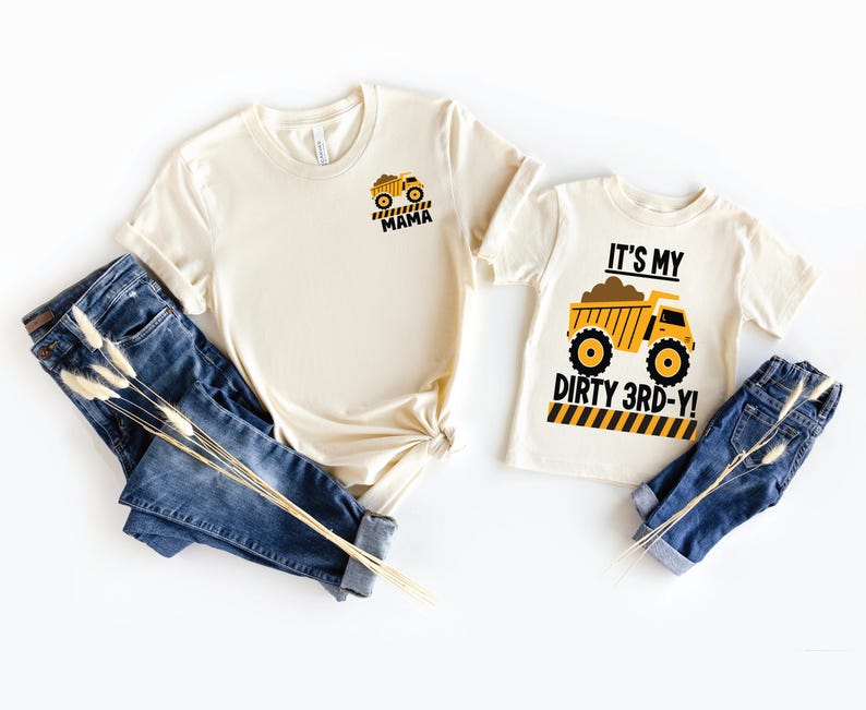 May include: Two cream-colored short-sleeve t-shirts and two pairs of blue denim jeans. One shirt features a small yellow dump truck graphic with the text MAMA. The other shirt features a large yellow dump truck graphic with the text IT'S MY DIRTY 3RD-Y!