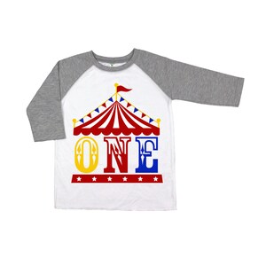 Puede incluir: Una camiseta de b&eacute;isbol blanca y gris con un gr&aacute;fico de carpa de circo rojo, blanco y azul y la palabra "ONE" en letras amarillas, azules y rojas. La camiseta tiene un patr&oacute;n de estrellas rojo, blanco y azul en la parte inferior.