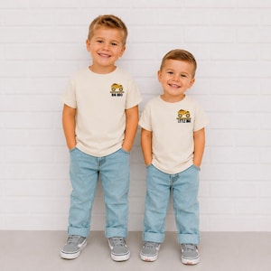 May include: Two cream-colored short-sleeve t-shirts with light blue jeans. The left shirt features a yellow construction truck graphic with the text BIG BRO. The right shirt features a yellow construction truck graphic with the text LITTLE BRO.