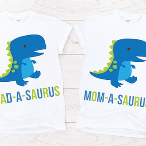 May include: Two white t-shirts with a blue dinosaur graphic and the text "DAD-A-SAURUS" and "MOM-A-SAURUS" printed on them.