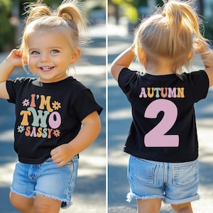 Może przedstawiać: Czarny t-shirt z kolorową grafiką, na kt&oacute;rej z przodu widnieje napis "I'm Two Sassy", a z tyłu "Autumn 2". Koszulkę nosi małe dziecko.