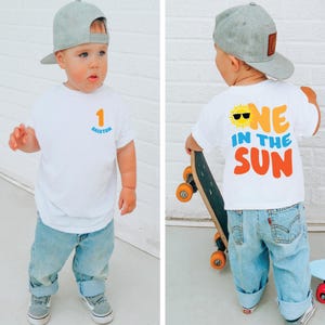 May include: A white short-sleeve t-shirt featuring a small orange number 1 and the name Brixton in blue on the front. The back displays a yellow sun graphic with black sunglasses and the text ONE IN THE SUN in orange and blue lettering.