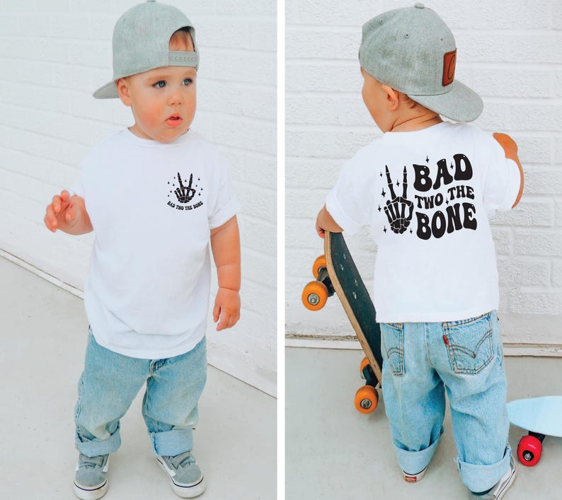 Puede incluir: Una camiseta blanca con una mano de esqueleto negra haciendo un signo de paz y el texto "Bad Two The Bone" en la parte delantera. La parte posterior de la camiseta tiene el texto "Bad Two The Bone" en una fuente retro. Un ni&ntilde;o peque&ntilde;o lleva la camiseta y una gorra de b&eacute;isbol gris. El ni&ntilde;o sostiene una patineta.