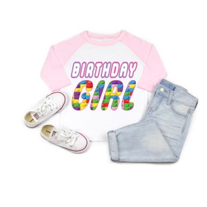 May include: White and pink baseball tee shirt with the text "Birthday Girl" in rainbow colours. The letters are made up of colourful Lego blocks.