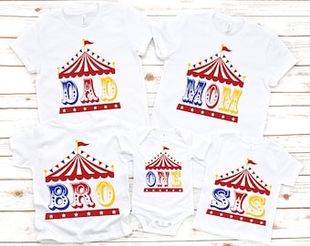 Puede incluir: Un conjunto de cinco camisetas blancas con un diseño de carpa de circo y las palabras "DAD", "MOM", "BRO", "ONE" y "SIS" en letras coloridas de estilo circense. Las camisetas son perfectas para un atuendo familiar a juego.