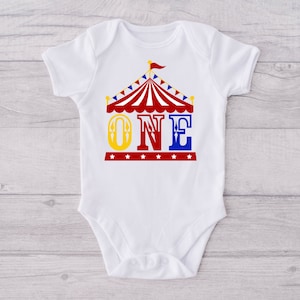 Puede incluir: Un body blanco para beb&eacute; con un gr&aacute;fico de carpa de circo roja, amarilla y azul y la palabra "ONE" en letras amarillas, rojas y azules.