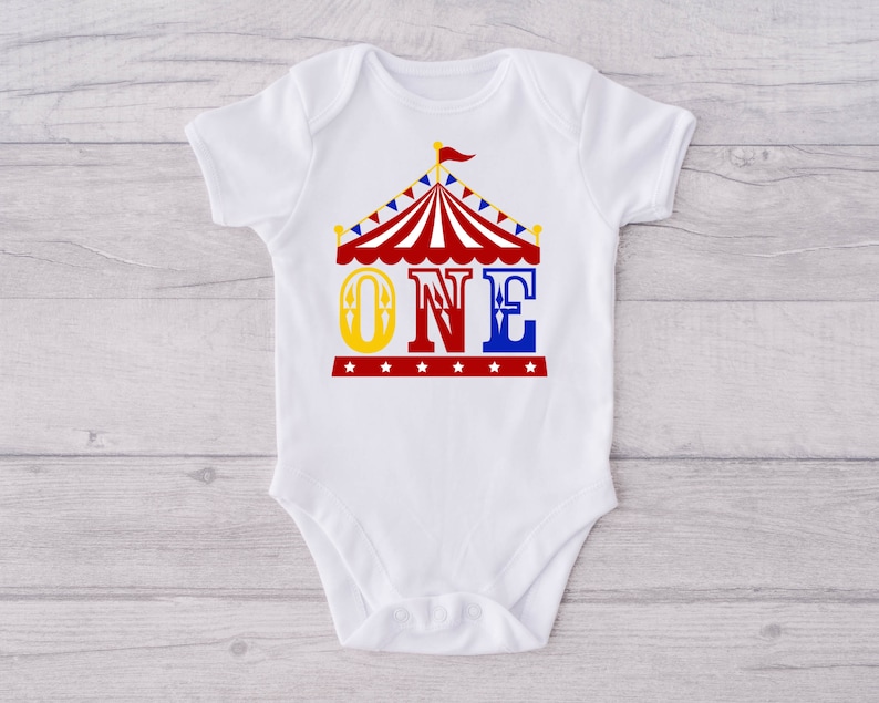 Puede incluir: Un body blanco para beb&eacute; con un gr&aacute;fico de carpa de circo roja, amarilla y azul y la palabra "ONE" en letras amarillas, rojas y azules.