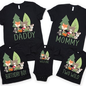 May include: A set of four black t-shirts with a woodland animal theme. The shirts feature a fox, bear, raccoon, and deer in a forest setting. The text on the shirts reads "Daddy", "Mommy", "Birthday Boy", and "Two Wild".