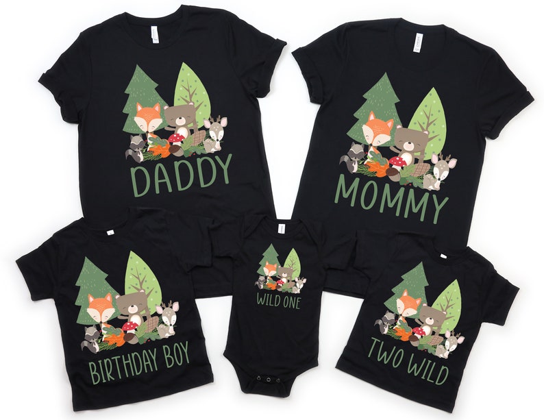 May include: A set of four black t-shirts with a woodland animal theme. The shirts feature a fox, bear, raccoon, and deer in a forest setting. The text on the shirts reads "Daddy", "Mommy", "Birthday Boy", and "Two Wild".