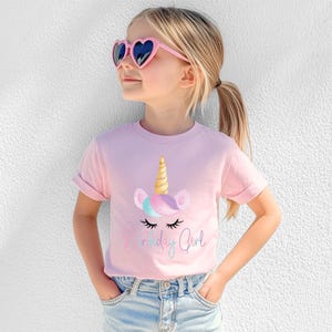 May include: A light pink t-shirt featuring a unicorn graphic with a golden horn, pastel mane, and the text "Birthday Girl". The shirt is worn by a young person wearing heart-shaped sunglasses.