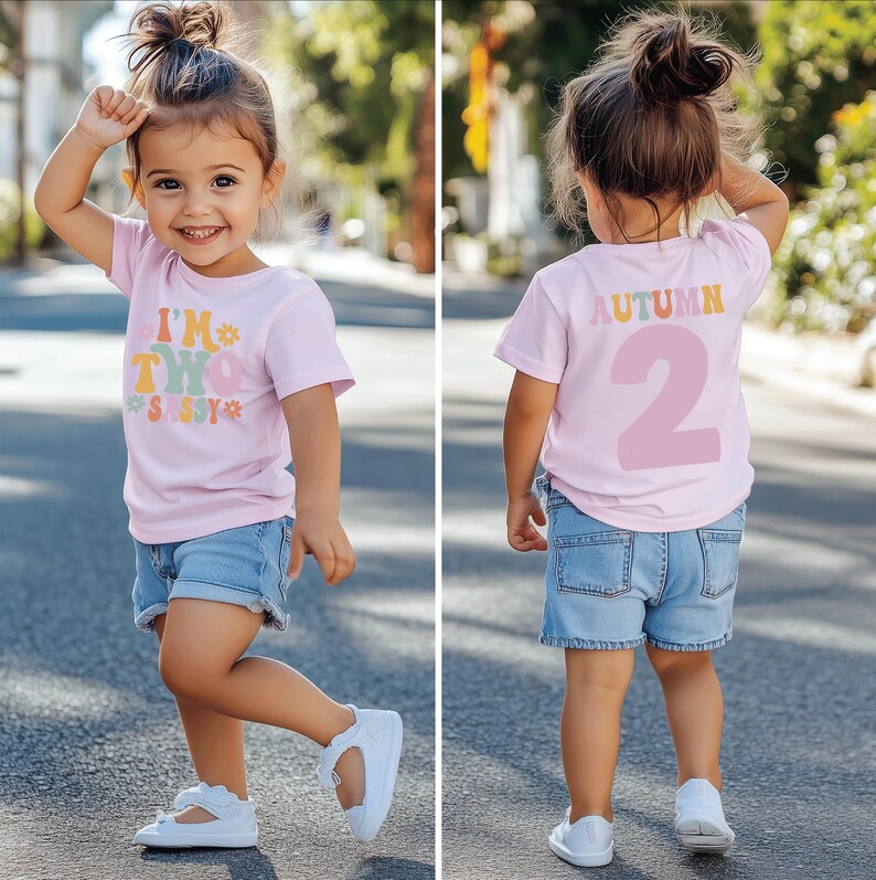 Może przedstawiać: R&oacute;żowa koszulka z napisem "I'm Two Sassy" z przodu i "Autumn 2" z tyłu. Koszulkę nosi dziecko.