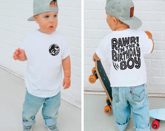 May include: A white t-shirt with a black dinosaur graphic on the front and the text "RAWR! I'M THE BIRTHDAY BOY" on the back. The shirt is being worn by a child wearing a grey baseball cap and blue jeans.