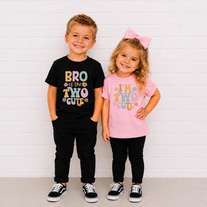 May include: A black t-shirt with the text BRO of the TWO CUTE and a pink t-shirt with the text I'M TWO CUTE, both featuring colorful retro-style lettering and flower graphics. The black shirt is paired with black pants, and the pink shirt is paired with black pants and a pink hair bow.